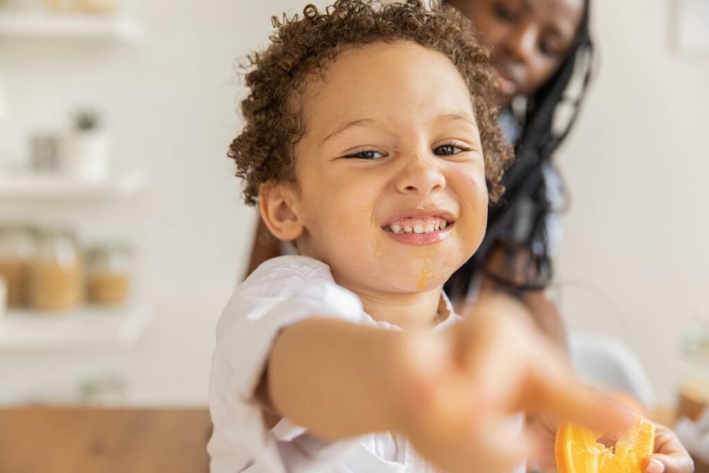 Joyful child with curly hair and orange slice playfully smiling indoors.