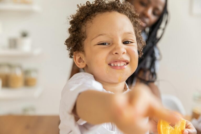 Joyful child with curly hair and orange slice playfully smiling indoors.